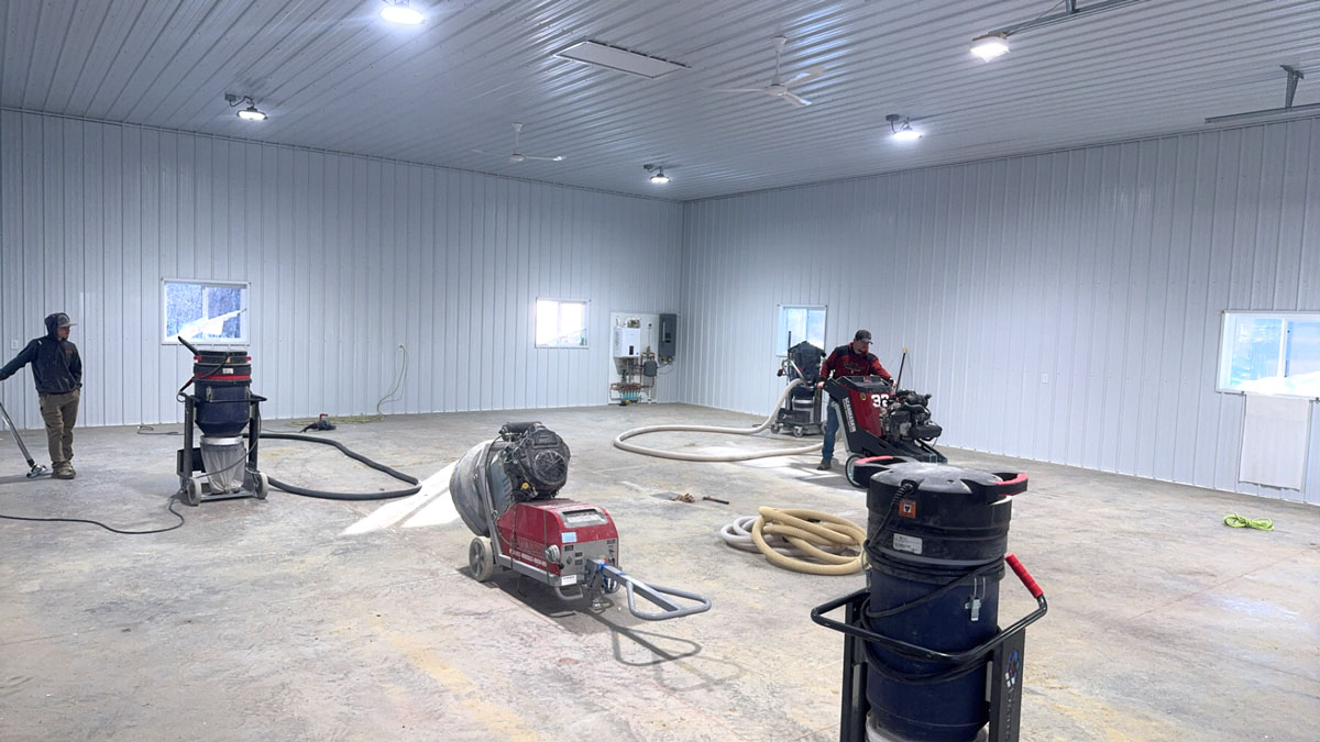 Two-man crew grinding and prepping a large metal-building floor with grinders and dust-collection vacuums